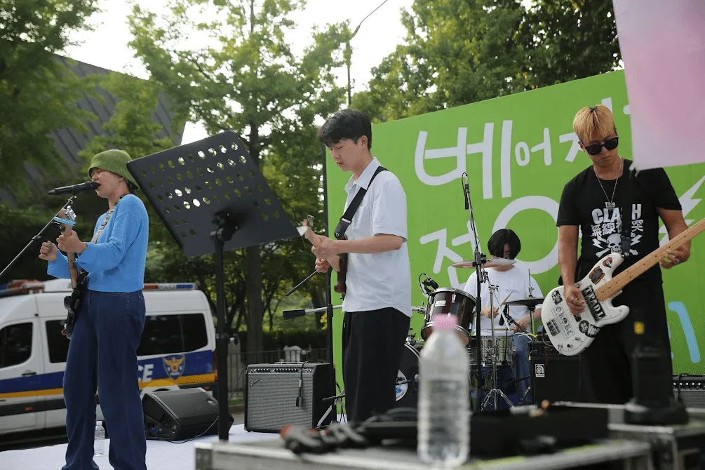 해질 무렵 초록 백드롭 앞 네 사람이 기타와 베이스, 드럼으로 연주하고 있다. 무대 옆에는 경찰 차량이 서 있다
